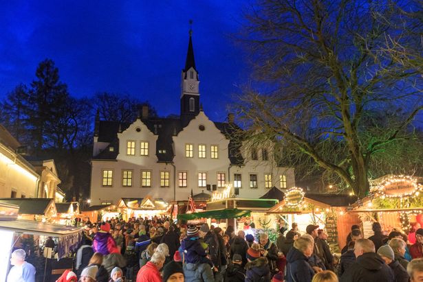 Christmas market with many ornate and decorated stalls. A large white building can be seen in the background - lined with white turrets and bay windows.