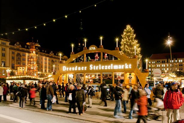 A Christmas market with a larger-than-life candle arch in the center with the words "Dresdner Striezelmarkt" can be seen. Numerous visitors walk through the Christmas market. The show booths and stalls are illuminated by attached Christmas decorations.