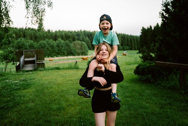 Große Schwester trägt kleinen Bruder auf den Schultern und läuft dabei über eine Wiese auf dem Bauernhof Kleines Vorwerk